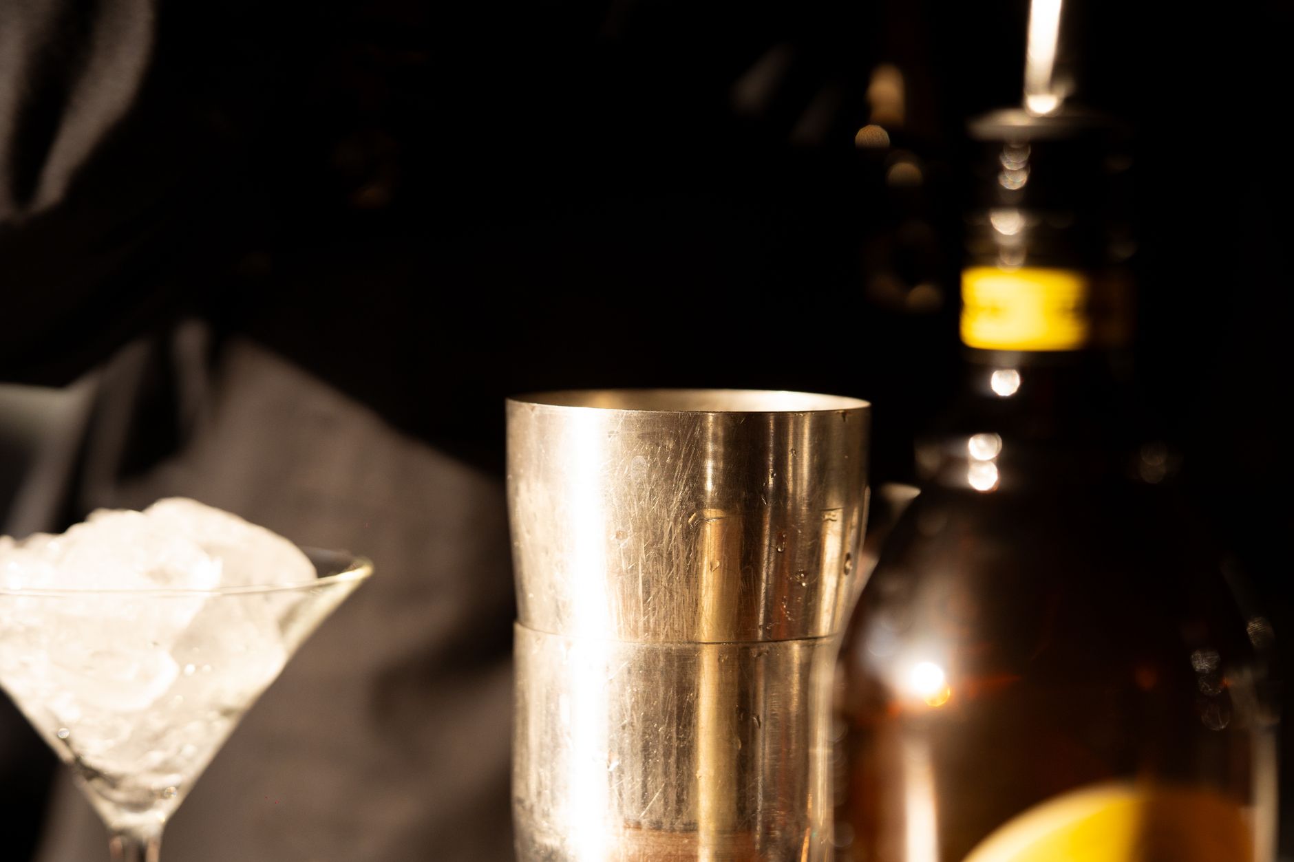 Pirinç shaker tini; solunda buz dolu martini kadehi, sağında odak dışı şişeler ve sarı etiket