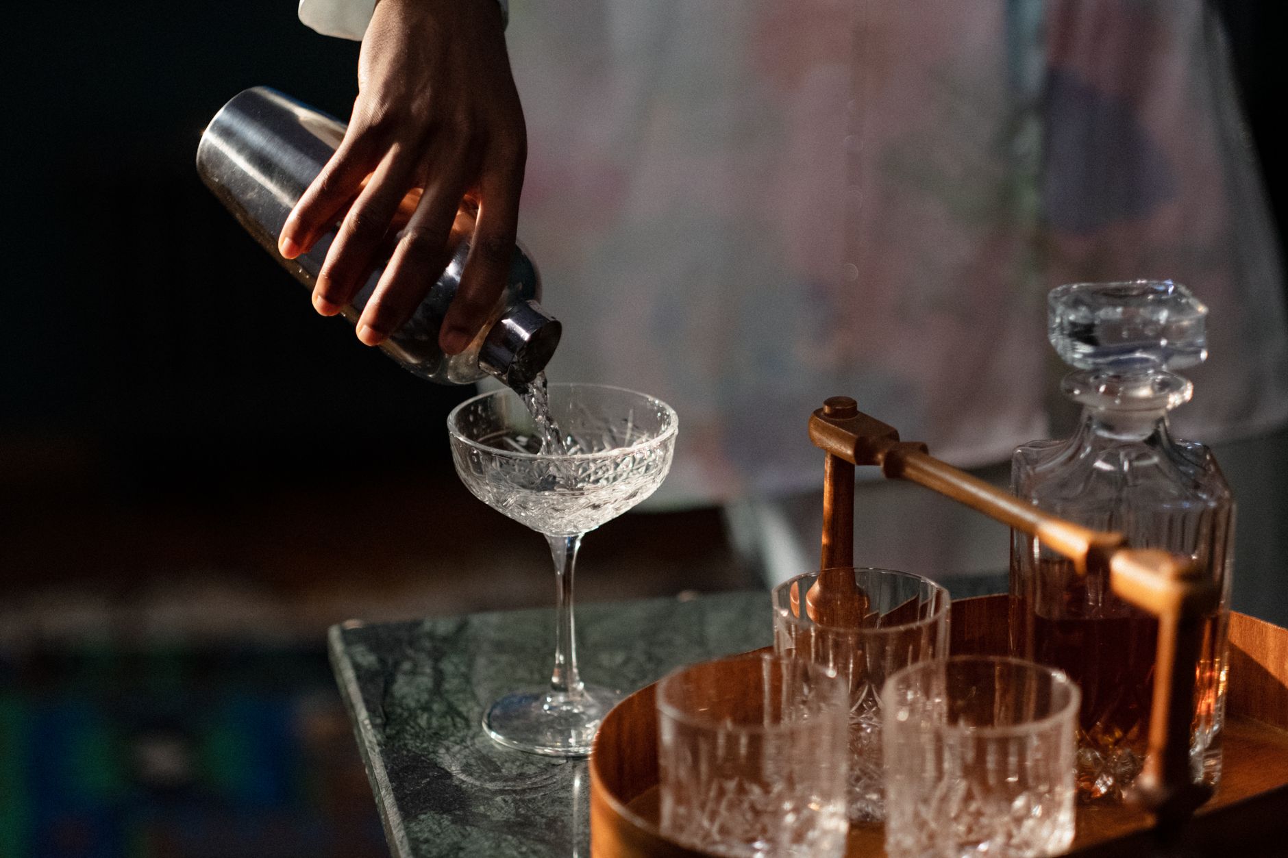 Barmenin eli shaker'dan kesme kristal kadehe süzüyor; yanda ahşap tepside dekanter ve bardaklar