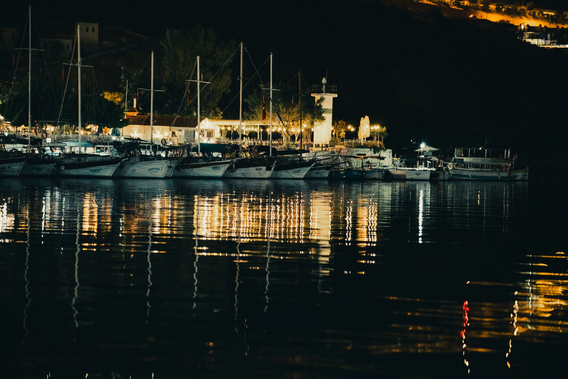 Gece marinası: bağlı tekneler ve kıyıda ışıkları yanmış teras, suya vuran yansımalar